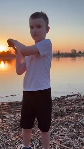 A young boy stands on the bank of a lake, a pile of reeds at his feet. He is wearing a white t-shirt and black shorts. Behind him, the setting sun casts an orange glow on the water and sky. He examines a small object in his hand, then picks up a twig and brandishes it. He leans forward and appears to throw something into the water.