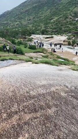The video shows a group of people gathered on a rocky landscape, possibly a riverbed or a hiking trail, surrounded by green hills. The camera is positioned on a higher rock surface, providing a view of the people below. Some individuals are standing and walking around, while others are sitting and relaxing. The environment consists of rocky terrain, patches of green grass, and bodies of water. The atmosphere appears to be casual and leisurely, suggesting a group outing or excursion in a natural setting.