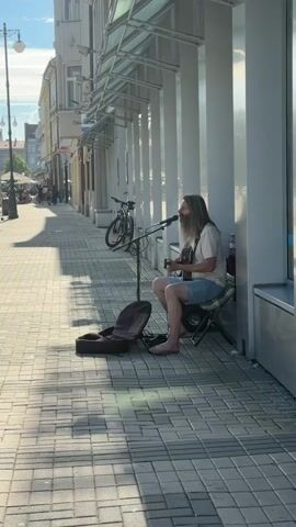 A man with long hair and a beard is sitting on a folding chair on a paved sidewalk in front of a building. He is playing an acoustic guitar and singing into a microphone. A guitar case is open on the ground next to him. There is a bicycle parked behind him. The scene appears to be in a city or town, with buildings and a street visible in the background. The sun is shining, casting shadows on the ground.