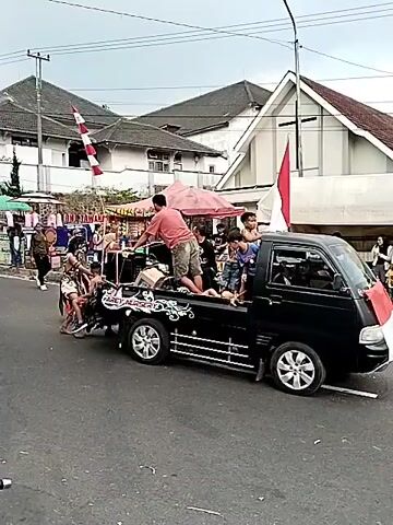 This is a video of a parade or cultural event taking place on a street. A black pickup truck is driving down the street, carrying several people, including children, in the bed of the truck. The truck is decorated with silver trim and has a speaker system. A man in a red shirt is standing in the back of the truck, holding onto something.

On the side of the street, there are stalls or vendors set up with tents. A group of people are walking in the parade, dressed in elaborate costumes that appear to be tribal or traditional. They are wearing headdresses, face paint, and outfits made of natural materials like leaves and animal skins. Some of them are carrying props, such as spears or shields.

On the sidewalk, there are spectators watching the parade. One person is holding up a smartphone to record the event. Indonesian flags are visible, suggesting the event may be related to a national holiday or celebration. The buildings in the background appear to be houses or businesses, with tiled roofs. The sky is overcast, indicating a cloudy day. The overall atmosphere seems festive and communal.