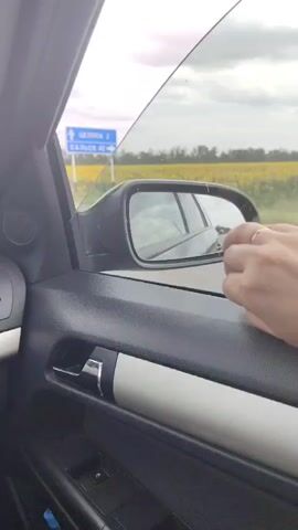 A hand with a ring is resting on the open window of a car. The car is driving past a field of sunflowers and a road sign that says "Ts 22 Sal'sk 420 km". The sky is cloudy.