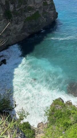 The video showcases a high-angle view of a coastal inlet. The turquoise waters churn and foam where waves crash against the rocky shore. A rugged cliff face rises on the left side of the frame, partly casting a shadow on the water below. Green vegetation clings to the rocks and the edge of the frame, adding a natural touch to the scene. The overall impression is one of natural beauty and the power of the ocean.