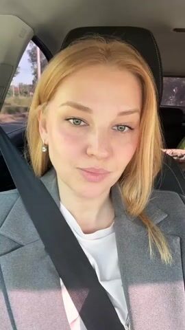 A blonde woman in a gray blazer and white shirt is in the driver's seat of a car, looking at the camera and smiling. The seatbelt is fastened across her chest. The interior of the car is visible, as are trees and sky through the windows. The video consists of short clips of the woman making faces and smiling.