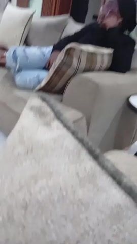 A person is resting on a beige sofa in what appears to be a living room. The person is wearing a black top, ripped light blue jeans, glasses, and a black head covering. They are propped up with a plaid pillow, and there are several other pillows on the sofa. The person appears to be looking off to the side with their hand near their face. The room has wooden trim around a doorway, and there is a small white table visible near the edge of the sofa.