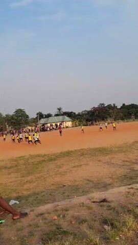 The video shows a dirt field where a soccer game is in progress. Numerous players in yellow and white jerseys are running around the field. A small crowd of people stands along the sidelines, watching the game. In the background, there is a building with a silver roof, surrounded by trees and vegetation. In the foreground, a person wearing black shorts and a black shirt is sitting on the ground, observing the game. The sky is clear and blue.