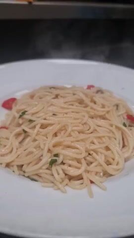 A close-up shot shows a white plate filled with spaghetti. The spaghetti is topped with chopped parsley and red pepper flakes. Steam rises from the pasta, indicating it is hot. The background is blurred, but appears to be a kitchen environment with dark countertops and a hint of orange in the upper left corner. The video focuses on the act of garnishing the pasta with fresh herbs.