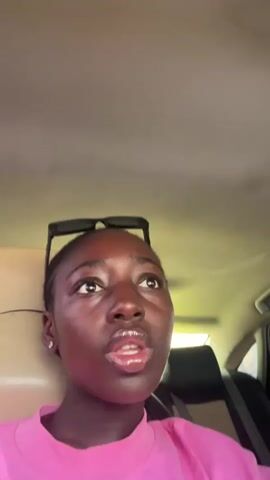 A young woman with dark skin and short hair, wearing a pink t-shirt and sunglasses on her head, is in the passenger seat of a car. Her head is turned slightly to the left as she looks up and to the side. She then turns her head and looks directly at the camera, speaking and making kissing gestures. The interior of the car is visible, including the headrest of the seat in front of her, and the ceiling of the car. The lighting suggests it is daytime.