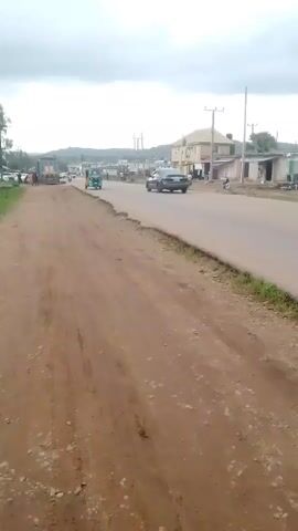 This media file captures a scene from Unguwar Saya, Nigeria, on a cloudy day, likely in the late morning or early afternoon. The foreground is dominated by a dirt road, showing tire tracks and loose gravel, indicating it's a less-developed area or a side road.

The main focus shifts to a paved road running parallel to the dirt path. Several vehicles are visible on this paved road, including a green and yellow auto-rickshaw (also known as a Keke Napep), a dark sedan, and several motorcycles. People are present on and around the road; some are riding the motorcycles, while others appear to be pedestrians or vendors near buildings.

The setting features a few buildings lining the paved road, suggesting a commercial or residential area. One building, in particular, is a multi-story structure with a light-colored facade and visible windows. Utility poles with power lines are scattered along the roadside, common in many populated areas. In the background, a line of hills or mountains can be seen under the overcast sky, adding depth to the landscape.

The activity appears to be the normal daily traffic and life in this part of Nigeria. The cloudy weather suggests it might be a humid day, typical of the region. There are no strong emotional cues conveyed; the scene is one of everyday activity. Some text is visible on the side of the auto-rickshaw, but it is too blurry to be deciphered. The overall impression is a glimpse into a typical roadside scene in a Nigerian town.