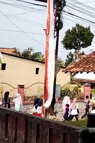 A group of children, some wearing hijabs, walk along a path next to a wooden fence. A tall flagpole with a red and white Indonesian flag is prominent in the foreground, leaning against a utility pole with wires. Behind the children, there is a building with beige walls and a tiled roof, and a decorative fence. A tree with green leaves is visible behind the building. The sky is overcast.