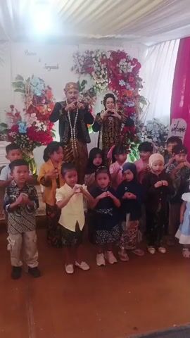 The video opens with a medium shot of a wedding reception, likely in a tent or indoor venue, decorated with white drapery and a floral arch. A couple, dressed in traditional attire, stands center frame behind a group of children. The groom wears a dark, embroidered jacket and pants with a headdress. The bride is in a patterned dress and headscarf. The children, ranging in age from approximately four to ten years old, are gathered in front of the couple. They are also dressed in various outfits, some traditional and some more casual. The children are either standing or sitting, and many appear to be engaged in a coordinated clapping or hand-waving activity. The backdrop includes a banner with text that reads "Paiman & Suryanah Welcome". To the right, a pink curtain hangs, and further back, more floral decorations are visible. The lighting is bright, suggesting an indoor event. The video appears to be a recording of a celebratory moment, possibly a children's performance or a family photo opportunity.