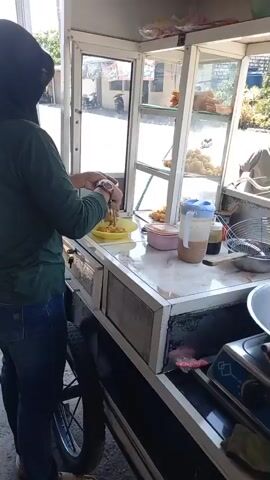 A person wearing a headscarf and jeans is standing behind a food cart, preparing food. The cart has a glass display case with various food items. A young boy on a bicycle is visible in the background. The scene appears to be outdoors, possibly on a street, with a road and buildings visible. A stove and cooking utensils are also part of the food cart setup.