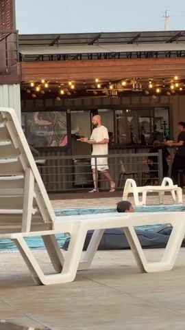 A man with a shaved head walks with a plate of food towards a building with large windows and string lights. He is wearing a white t-shirt and patterned shorts. To his right, another man in a dark shirt walks away from the building. In the foreground, a white lounge chair sits next to a swimming pool. A child lies on a beanbag chair by the pool. A clear beach ball with colorful shapes is partially submerged in the water.