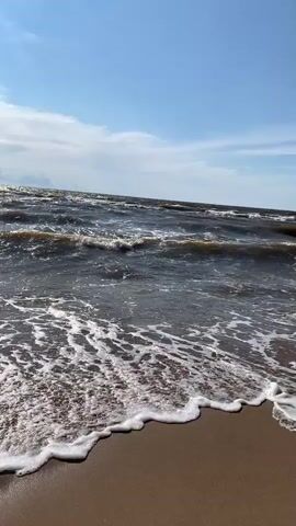 The video shows a beach with waves crashing on the shore. The water is dark and choppy, with white foam washing up on the sand. The sky is blue with some clouds. There are a few people visible in the distance swimming in the ocean and walking along the beach. The camera pans across the scene, capturing the movement of the waves and the expanse of the ocean.