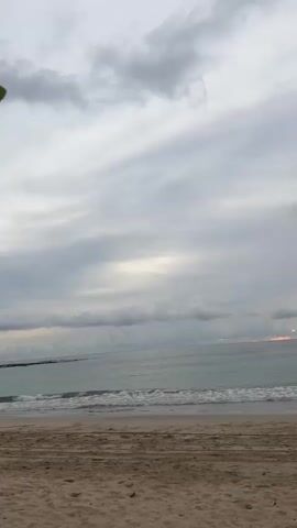 A static, eye-level shot of a beach at dusk. The sand in the foreground is a light brown, with faint tracks and imprints. Beyond the sand, the ocean is a muted turquoise, with small waves breaking gently on the shore. The horizon line is visible, separating the sea from the sky, which is filled with a heavy blanket of grey clouds. In the distance, on the right side of the frame, a dark, mountainous landmass juts out into the sea. The sky above the horizon is illuminated with faint hues of orange and yellow, suggesting the setting sun may be partially visible behind the clouds or landmass. There are no people or animals visible in the frame. The overall mood is serene and tranquil.