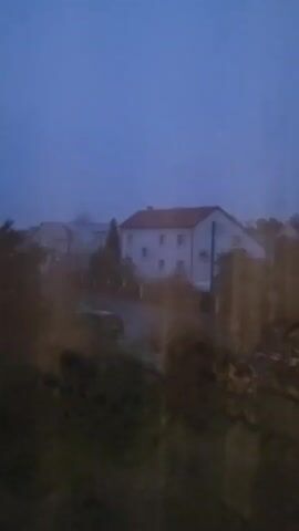 A video shows a hazy view of houses and trees under a dusky sky. The foreground is blurred, suggesting the footage was captured through a window or dirty lens.