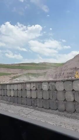 The video is a first-person view from a vehicle traveling along a paved road. A retaining wall constructed of interlocking concrete blocks runs along the right side of the road. Each block in the wall appears to have a white sign or decal affixed to its face, some with images or text that are not clearly discernible. Behind the retaining wall, the terrain slopes upward, featuring terraced hillsides covered in patchy green vegetation and exposed earth. The sky above is blue with scattered white clouds, indicating daylight. The overall scene suggests a landscape undergoing development or stabilization, possibly a construction site or a mountainous region with infrastructure.