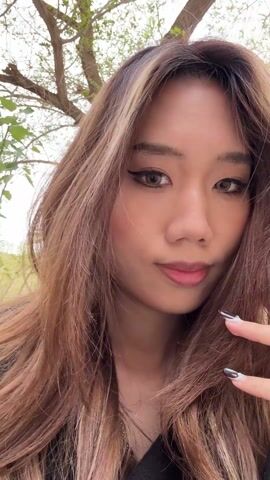 A young woman with long brown hair and a light blonde highlight near her face is standing outdoors. She looks directly at the camera with a slight smile. In the background are trees and what appears to be a stone wall or structure. The sky is overcast. The woman is wearing a black top and her nails have a light-colored design. The video is a close-up shot focusing on her face and upper body. The camera slightly moves around her face, with the background changing around her hair.