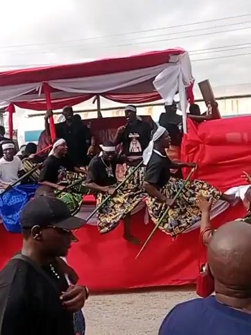 The video shows a group of people on a red and white float, possibly a parade or a celebratory event. Several individuals on the float are wearing traditional attire, including patterned skirts and headbands, and are holding long poles. Some are seated, while others stand. In the foreground and around the float, more people are visible, some of whom are interacting with those on the float. The environment appears to be outdoors, with buildings and a cloudy sky in the background. There are also some vehicles, including a motorcycle and what appears to be a three-wheeled vehicle, suggesting a street setting. The overall atmosphere seems lively and festive.
