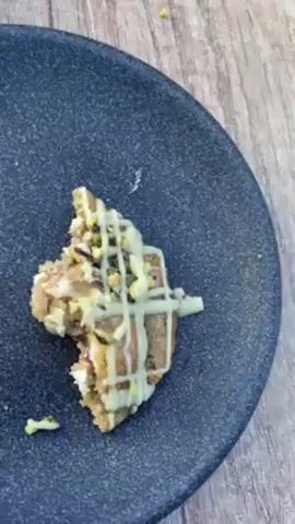 The video shows plates with dessert on a table outside. There are two plates with broken pieces of cookies on them, each drizzled with a light-colored icing and sprinkled with green nuts, and a cup of coffee with milk foam art on top. The table is outdoors, with a wooden top and a brown serving tray nearby. The camera pulls back to reveal other tables and chairs, suggesting an outdoor café. The tables are metal with green metal chairs. The background shows a pedestrian street with buildings on either side, indicating a town or city setting. Some people are seen walking along the street. The sky is visible with some light clouds.