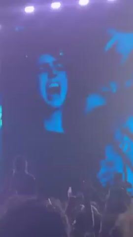 The video appears to be taken at a concert. In the first frame, a large screen displays a woman singing with her mouth open. The scene then quickly transitions to darkness before brightening again to reveal a crowded audience with bright stage lights and a large screen displaying abstract blue graphics. The video ends with a view of the stage, barely visible through a haze of light.