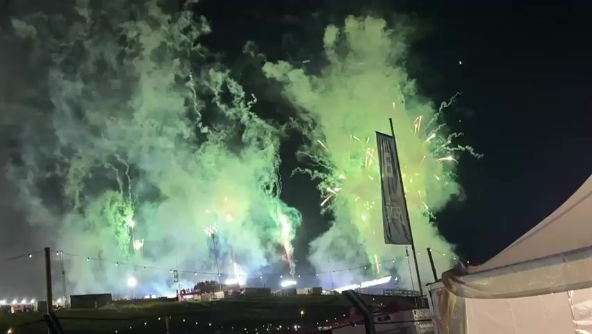The video captures a nighttime fireworks display from a slightly elevated, stationary perspective. Numerous green and yellow fireworks explode, filling the dark sky with large plumes of light green and white smoke. Several trails of light ascend from the ground, indicating active launches. A dark blue flag with white lettering, partially obscured by a white tent structure on the right, stands prominently in the foreground. In the distant background, various small lights illuminate the landscape, suggesting a large outdoor event or festival area. The overall scene is dynamic with continuous explosions and smoke effects.