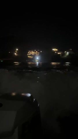 The video depicts a nighttime scene on a body of water. The perspective appears to be from a moving boat.  Across the water, lights from buildings or structures can be seen along the shoreline.  The water is turbulent and agitated, with white foam visible. The only objects visible in the foreground appear to be parts of the boat. The lighting is poor overall and the scene is dark, with the lights in the distance providing the only significant illumination.