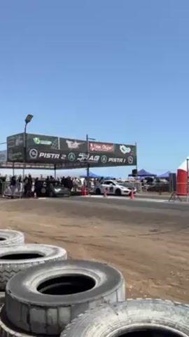 The video shows a drag race event. There are two cars at the starting line of a drag strip. There is a black car on the left and a white car on the right. A crowd of people is standing behind a barrier near the starting line. Tires are stacked in the foreground. There is a banner with the words "Pista 2" and "Pista 1". The sky is clear and blue.
