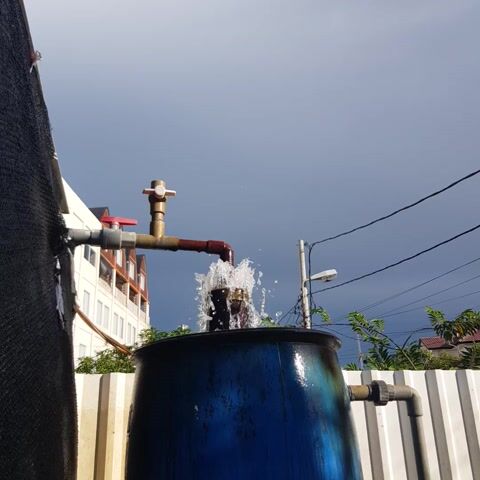 This is a close-up, low-angle shot of a blue water tank from below, with the sky as the background. A red pipe with a valve is attached to the tank, and water is spraying out of the valve, creating a splash. The scene is outdoors, with some greenery and a building visible in the distance. The video appears to be shot on a sunny day, as indicated by the bright blue sky. The camera is slightly tilted, giving the image a dynamic feel. There are no people visible in the frame. The focus is on the water spraying from the valve.