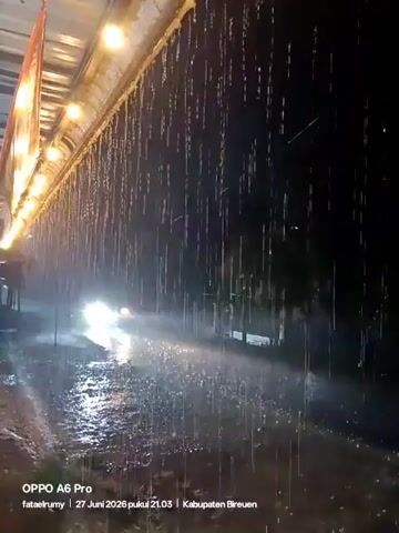 A night scene shows heavy rain falling from an overhang illuminated by several lights. The street below is wet and reflective, with water pooling in places. Cars with headlights on are driving through the rain, their lights illuminating the spray. One person is walking or riding a motorcycle on the left side of the street. The timestamp in the bottom corner indicates the video was recorded on June 27, 2026, at 9:03 PM, in Kabupaten Bireuen.