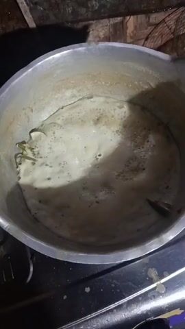 The video shows a close-up, overhead shot of a pot of food bubbling on a stove. The pot is made of aluminum and has some residue on the sides. The food inside appears to be a light-colored liquid with a foamy texture, and there are some green leafy ingredients and what looks like a piece of fish or meat submerged in the liquid. The stove is black and has a metallic surface. The background is dimly lit and appears to be a rustic kitchen setting with wooden walls and possibly a fan. The lighting suggests it is nighttime or the room is poorly lit. The focus is entirely on the pot and its contents.