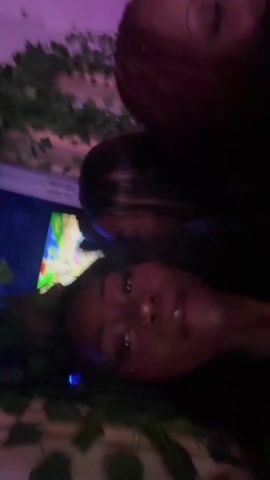 A dark-skinned woman with long, dark hair smiles and sings along to music. She is lying down and faces the camera directly. There are decorations, including artificial vines, in the background. There is also a lit screen visible, and a purple light fills the scene.