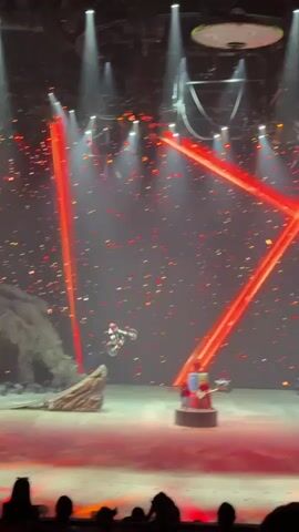 The video shows a stage production with a red and black color scheme. A motorcyclist performs a jump through the air, surrounded by red laser beams and light effects. After the jump, the motorcyclist rides on one wheel. In the background, performers in white and red outfits wave checkered flags. The stage includes rock-like props, a structure resembling a gate, and other stage elements. There are bright spotlights shining down on the stage. The audience is visible at the bottom of the frame.