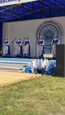 Five women in traditional blue and white clothing stand on a stage holding white fabric with blue stripes. They are positioned behind music stands. The stage is covered by a blue tent structure. A large circular banner is visible behind the women. In front of the stage, several small white flags on blue poles are arranged. The ground in the foreground is covered with green grass and a wooden structure. The overall scene appears to be an outdoor performance or festival.