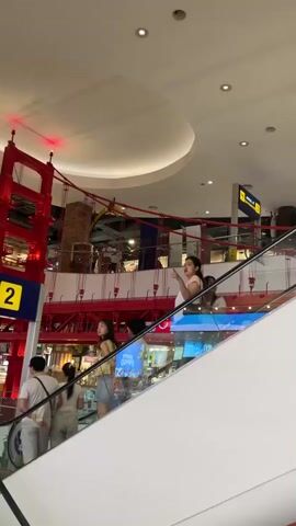 The video shows a multi-level indoor space resembling a shopping mall with escalators and decorative elements. On the left side of the frame, a red structure resembling a suspension bridge is visible, with a sign that says "ARRIVAL AT TOKYO" and the number "2." Several people are seen on the escalators, moving in different directions. On the lower escalator, a man in a green t-shirt and shorts walks down, holding an ice cream cone. A woman in a black dress is on the same escalator, looking to her left. On the upper escalator, a woman in a white top and blue jeans is standing, looking forward. Another woman in a yellow top and denim shorts is behind her, holding a bag. The environment appears bright and well-lit, with a large, curved ceiling.