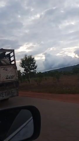 A person is standing on the back of a flatbed truck loaded with sacks. The truck has markings on the side, including the words "GO" and what appears to be a Nike logo. There is also writing on the tailgate that reads "OKADA TAKE TIME." The truck is in motion on a road with vegetation and hills in the background. The sky is overcast. The camera is positioned within another vehicle, as evidenced by the reflection in a side mirror.