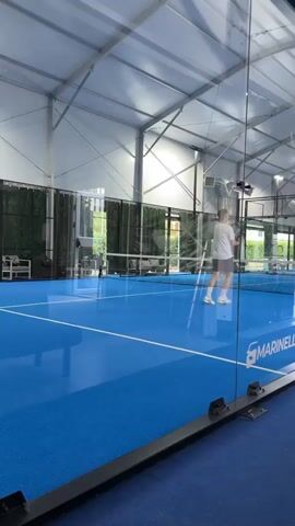 A young man in a white t-shirt and dark shorts is playing padel tennis indoors. He is on a blue court with glass walls. He is holding a padel racket and appears to be in the middle of a rally or preparing to hit the ball. The court is well-lit with artificial lighting from the ceiling. The background shows the structure of the indoor facility and glimpses of greenery outside through the glass. The man is the only person clearly visible on the court.