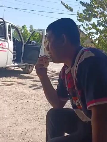 A man is sitting in the foreground, eating. A white pickup truck is parked to his left with its doors open. The truck has "KOPERASI DESA MERAH PUTIH" written on its side, along with the number "4x4". The truck bed is filled with large white plastic containers. The scene appears to be outdoors on a sunny day, with a dirt or gravel ground. In the background, there are trees and buildings. The camera pans from the man to the truck.
