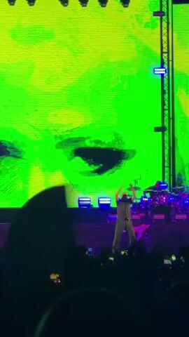 A concert performance is captured from the perspective of the audience. The stage features a large screen displaying abstract, colorful visuals, primarily green and red. A performer, dressed in dark clothing, is visible center-stage, raising their arms and seemingly interacting with the crowd. To the right of the stage, a drum set is partially visible. The audience, silhouetted in the foreground, is holding up phones, suggesting they are recording the event. The lighting is dynamic, with blue and purple lights illuminating the stage and the screen's colors shifting. The overall atmosphere is energetic, typical of a live music event.