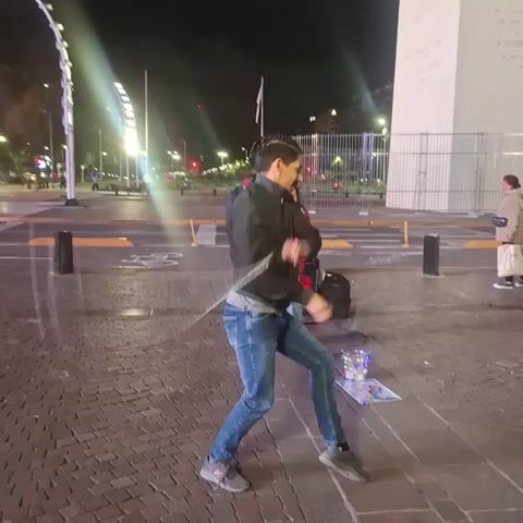 At night, on a pedestrian walkway made of cobblestones near a street, a man is dancing. He's wearing a dark jacket over a gray shirt, blue jeans, and gray sneakers. He is facing the camera and moving his arms and legs. To his right, a young girl in a red jacket is singing into a microphone. A speaker and other equipment are on the ground behind her. Behind them is a street with cars passing. In the background is a large white monument, possibly an obelisk. Another pedestrian walks by on the right side of the frame, carrying bags. Streetlights and building lights illuminate the scene.