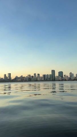 The video shows a skyline of tall buildings at sunset across a body of water. The sky is blue fading to yellow on the horizon. The water is calm with ripples and reflects the colors of the sky and the buildings. No people are visible.