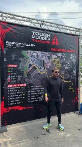 The video shows a man standing in front of a "Tough Mudder Thailand" banner, posing for the camera. The man is wearing sunglasses, a long-sleeved black shirt, black shorts, black leggings, and athletic shoes. He is holding a gray bag over his left shoulder. The banner behind him shows a course map with a legend and an obstacle menu. The event takes place in Wisdom Valley, Pattaya, Thailand, in May of 2026. He makes a peace sign for the camera. There is a structure of scaffolding holding up the banner and music playing in the background. The weather appears to be overcast.