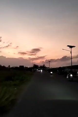 The video shows a view of a dark asphalt road stretching into the distance at dusk. The sky above is a blend of pale pink, orange, and grey hues, indicating the setting sun. Several scattered clouds partially obscure the sky. On the left side of the road, there's some greenery, while on the right, there are simple structures resembling covered market stalls or shops. In the distance, a few vehicles with their headlights on are visible. Two motorcycles are seen on the road, one closer to the camera and another further away. The camera is either stationary or moving very slowly, capturing the scene head-on. A solar-powered street light is visible on the right side.