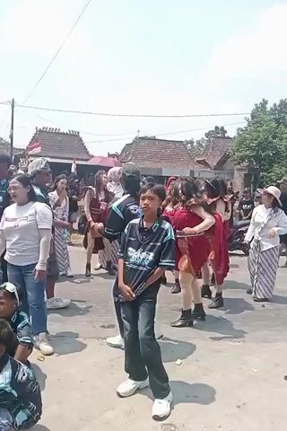 This is a video clip of an outdoor event in what appears to be a village setting. The scene is filled with people of various ages, some dressed in traditional clothing. In the foreground, a young boy in a blue and black patterned t-shirt and dark pants stands facing the camera. Behind him, a group of women in colorful dresses are dancing. To the right, a man in a black t-shirt and jeans is also dancing, facing away from the camera. In the background, there are houses with traditional tiled roofs and trees. The event appears to be a celebration or festival, possibly involving traditional dance. The lighting suggests it is daytime, with bright sunlight casting shadows.