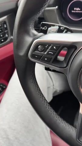 A close-up video shows the steering wheel of a Porsche car. The wheel is black leather with silver accents and control buttons on the left side. The Porsche emblem is in the center of the steering wheel. The dashboard is visible behind the steering wheel, showing gauges and a screen. The interior of the car includes red leather seats.