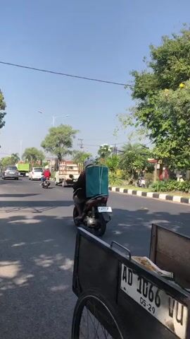 The video shows a person riding a three-wheeled bicycle cart down a street on a clear day. The cart has a black cargo box with a license plate that reads "AD 1066 VU". The rider is wearing a light brown jacket, dark pants, and a green hat. The street is paved and lined with trees and some buildings. There are other vehicles on the road, including cars, motorcycles, and trucks. The scene appears to be in an urban or suburban area. The video is shot from a slightly elevated perspective, looking down at the cart and the street. The sun is casting shadows on the road.