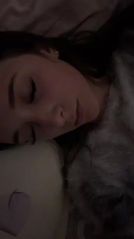 A young woman with long dark hair, wearing a light gray fuzzy top, lies on a white pillow with her eyes closed. She slowly opens her eyes and looks directly at the camera.