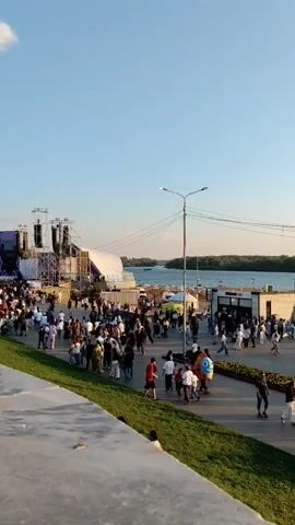 The video shows an outdoor gathering with many people walking around. There are several structures in the background, including what appears to be a stage or large event setup on the left, and several smaller booths or kiosks. A body of water, likely a river or lake, is visible behind the gathering. The sky is clear and blue, suggesting daytime. People are dressed in casual clothing, and some are pushing strollers or carrying bags. There is a grassy area in the foreground. The overall atmosphere appears to be a public event or festival.