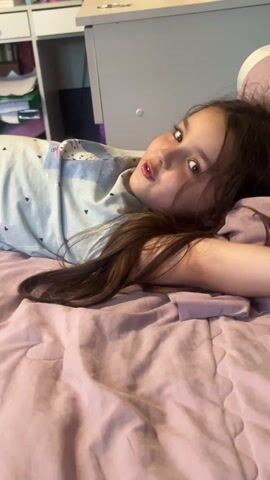 A young girl lies on a pink comforter on a bed, facing the camera. She has long, dark brown hair and is wearing a light blue t-shirt with a subtle pattern of small white shapes. Her head is resting on a pillow. To the left of the bed, a white desk with some papers and folders is visible. The lighting appears to be natural and soft.