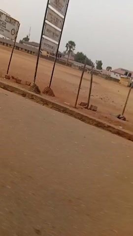 The video shows a roadside view in a rural area. A dirt field is visible, with a signpost near the road and a building in the background. The sky is overcast. A man walks across the foreground from left to right. The field transitions from bare dirt and debris to overgrown grass as the view moves further into the field. The road is made of a dark, possibly paved material, and appears to be slightly elevated in relation to the field.