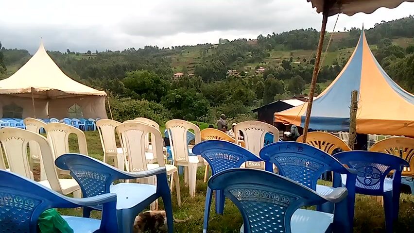 The video shows a rural area, likely in Africa, prepared for an outdoor event. A field is filled with rows of blue and white plastic chairs facing toward the camera. Two tents, one mostly white and the other a blue and orange mix, are visible. In the background, a hilly landscape covered with trees and scattered buildings is seen under a cloudy sky. A dog wanders among the chairs. Later in the video, a closer shot reveals the hillside landscape, showing trees, scattered houses, and open fields.